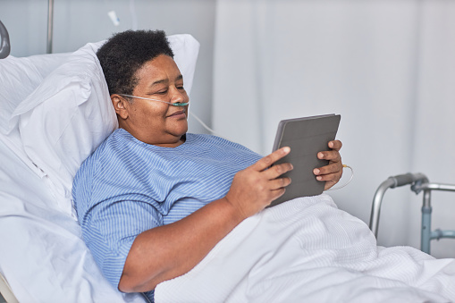 Side view portrait of African American senior woman using digital tablet while laying on bed in hospital