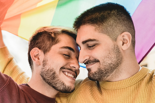 Happy gay men couple having tender moment under rainbow LGBT flag outdoor - Diversity love concept - Soft focus on right male face
