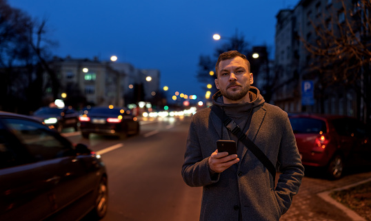 Adult man standing near the road in city holding phone in hand and looking away.