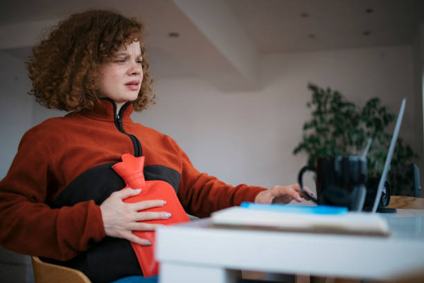 Young woman using a hot water bag for the stomach ache while working from home stock photo