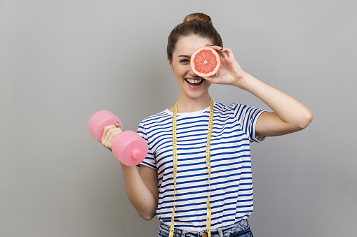 Woman with tape measure on shoulders holding dumbbell and covering eye with half of grapefruit, keeping diet to reduce extra centimeters on body. Indoor studio shot isolated on gray background.