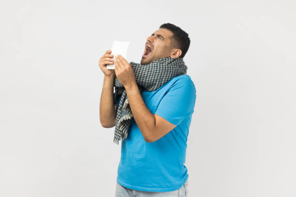 Sick man wrapped in warm scarf standing and sneezing, catching cold in winter, has high temperature. stock photo
