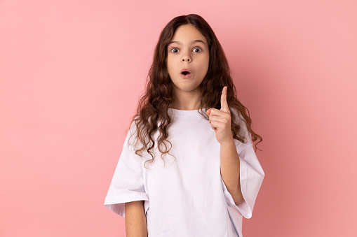 Portrait of surprised little girl wearing white T-shirt raising finger up, has sudden great idea, looking at camera with open mouth. Indoor studio shot isolated on pink background.