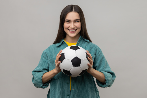 Pretty dark haired woman looking at camera with pleasant smile, holding soccer ball, watching football match, wearing casual style jacket. Indoor studio shot isolated on gray background.