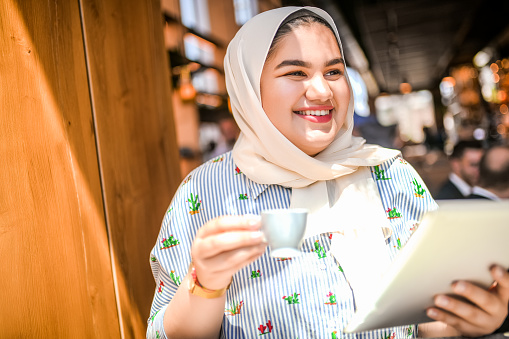 Smiling Muslim woman using digital tablet at the bar whole drinking a coffee