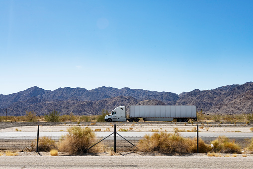 In Desert Center, United States a tractor trailer truck drives across the Mojave Desert hauling a cargo shipping container on a sunny spring day.