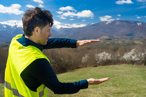 A man holding his hands out with palms facing up and down, wearing a yellow reflective jacket with distant mountains and woodland in the background