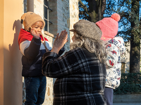 Portrait of african little kid in warm outerwear colorful vest and knit beanie cap with earflaps, and latino grandma hugging and playing