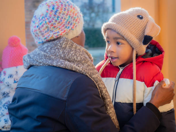 Grandma hugging caring smiling african american kid dressed warm with wool beanie knit cap with earflaps in fall winter outdoors at sunset Portrait of african little kid in warm outerwear colorful vest and knit beanie cap with earflaps, and mother caring and bonding half dozen babies stock pictures, royalty-free photos & images