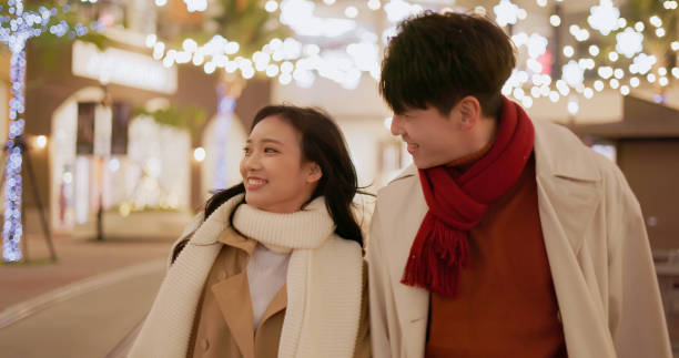 asian young couple wearing scarves with shopping bags are chatting and walking on light decorated street while dating in snowy winter day at night