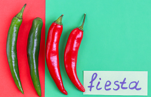 Four hot green and red chili peppers with the inscription on paper: fiesta and copy-space lie on a red-green background, flat close-up. Cinco de Mayo concept, minimalism, blanks.