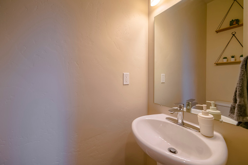 Small windowless bathroom with mirror and sink with white handwash bottle. Bathroom interior with light brown walls and a hanging shelves with succulents seen from the mirror reflection.