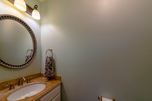 Windowless bathroom interior with gray walls and wall lighting fixtures. There is a single vanity sink on the left with round framed mirror and hanging brown towel on a ring holder on the left.