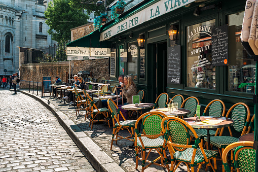 Paris, France - May 25, 2016: People drinking coffee in typical outdoor coffee shop with vintage chairs and tables on the sidewalk of the cobblestone street in Montmartre - famous and very popular neighborhood in Paris.