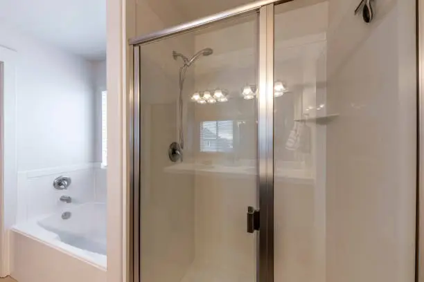 Shower stall with glass door and wall-mounted showerhead beside the bathtub with wall-mounted faucet. There is a bathtub on the left beside the shower stall on the right with reflection of the window.