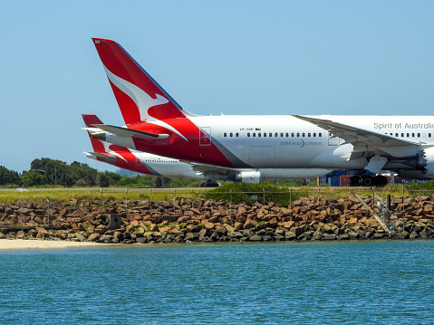 The vertical stabilisers and fuselages of two Qantas planes at Sydney Kingsford-Smith Airport.  Nearest is a Boeing 787-9, VH-ZNB, taxiing to the southern end of the runway before heading to Santiago as flight QF27.  In the distance is a Boeing 737-838, VH-VXG, flight number QF649, which is taxiing to the main north-south runway before taking off and heading to Perth.  This image was taken from the Botany side of the Cooks River, on a sunny afternoon on 26 February 2023.