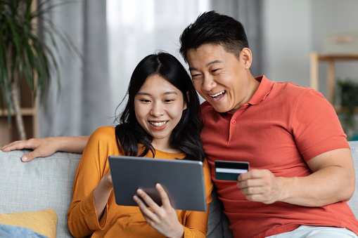 Excited asian husband and wife embracing, using digital tablet and plastic credit card, cheerful korean family paying for goods and services online while staying home, shopping on Internet, copy space