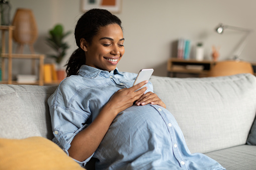 Happy Pregnant African American Woman Using Mobile Phone Texting Networking In Social Media Sitting On Couch At Home. Pregnancy Lifestyle, Gadgets And Communication Concept