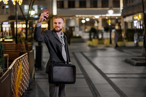 A handsome young man is walking in the city streets and taking a self-portrait photo over a smartphone during an autumn night in the city.