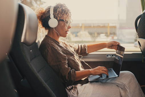 Mid age woman using computer inside transport bus vehicle during business or vacation travel. Modern work digital lifestyle. Female people with headphones enjoy transport. Traveler lady with laptop