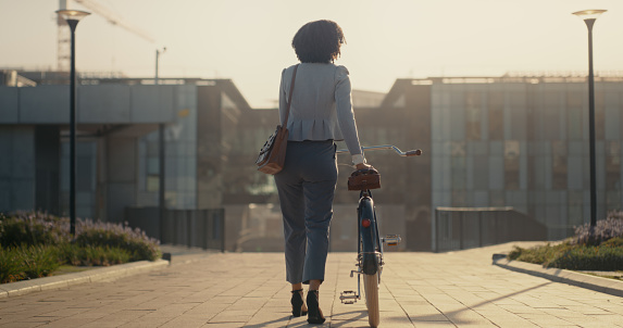 Bicycle, city and business black woman travel in street for eco friendly, sustainable or carbon footprint transport. Rear of professional person walking or cycling in morning sunrise on an urban road