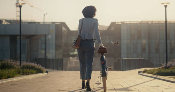 bicycle, city and business black woman travel in street for eco friendly, sustainable or carbon footprint transport. rear of professional person walking or cycling in morning sunrise on an urban road - sunrise city of sunrise street road imagens e fotografias de stock