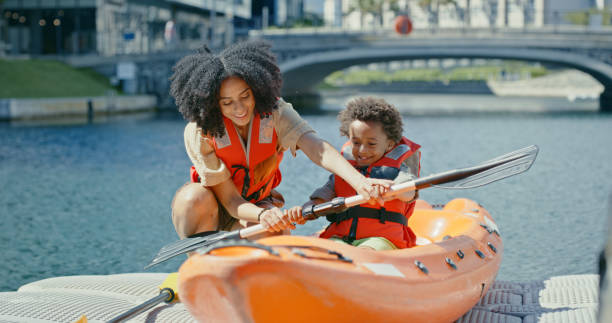 madre che insegna al bambino con pagaia, kayak e sport acquatici sul fiume o sul lago, tempo di vacanza in felice avventura estiva in svezia. viaggio, vacanza e donna nera con il figlio in canoa pagaiando nel canale della città - skiff time travel locations sports and fitness foto e immagini stock