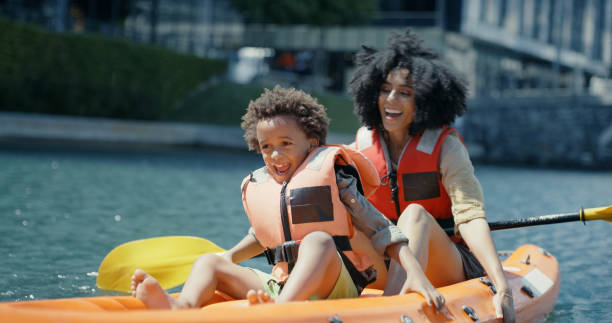 madre, niño y kayak en el río o lago, divertido tiempo de vacaciones con deportes acuáticos, verano y feliz aventura en suecia. viaje, vacaciones y mujer negra con hijo en canoa remando en el canal de la ciudad con sonrisa - skiff time travel locations sports and fitness fotografías e imágenes de stock