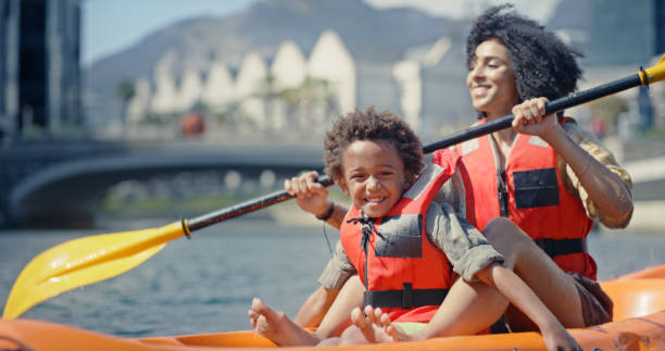 madre, bambino e kayak pagaiando sul fiume o sul lago, divertimento in vacanza con sport acquatici, estate e felice avventura. viaggio, vacanza e donna nera con figlio, canoa e pagaia nel canale della città con il sorriso - skiff time travel locations sports and fitness foto e immagini stock