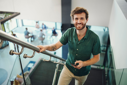 Agency, employee and portrait of man on office stairs in a company happy and smiling and holding a phone. Worker, intern or trainee excited with a positive mindset at the workplace by steps