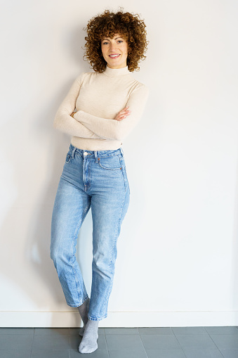 Full body of curly haired female in casual clothes leaning on white wall while standing with hands crossed looking at camera and smiling