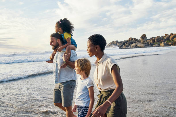 Family, kids and piggyback walking by sunset beach, nature or ocean in relax summer holiday, interracial vacation or freedom travel. Children, bonding and parents in shoulder carry trust by sea waves stock photo