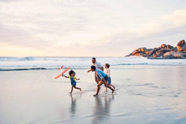 Happy family, running or flying toys on sunset beach or ocean in freedom holiday, energy bonding or travel playing fun. Sea, children or kids with airplane, interracial parents or aeroplane by nature stock photo