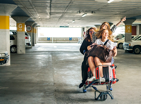 Laughing group of female friends riding on a luggage cart through an airport parking terminal before a flight