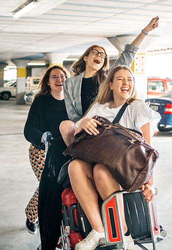 Group of laughing female friends riding on a luggage cart through an airport parking terminal before a flight