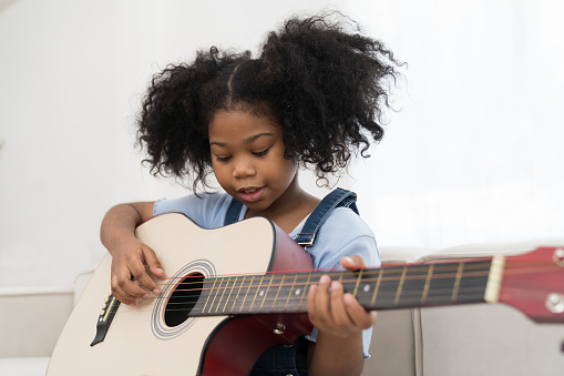 Happy afro girl playing and practice guitar. African American little child girl with curly hair playing guitar on sofa at home. Smiling child girl sitting on couch with guitar in living room