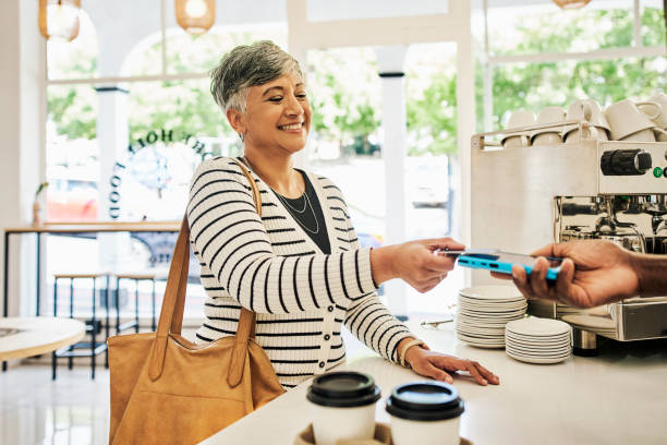 Payment, tapping and woman at a cafe for coffee or buying breakfast with a credit card. Retail, smile and mature customer paying for food or a drink on a machine with a server at a restaurant stock photo