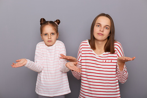 Portrait of uncertain woman and little girl with hair buns wearing casual clothes standing isolated over gray background, family posing with shrugging shoulders, having doubtful expression.