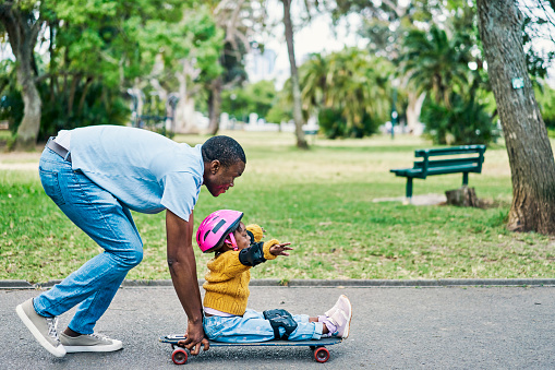 Field, skateboard and dad with his girl child teaching her to skate in an outdoor park. Sport, skating and African man helping his daughter or kid have fun in the road in nature or green garden.