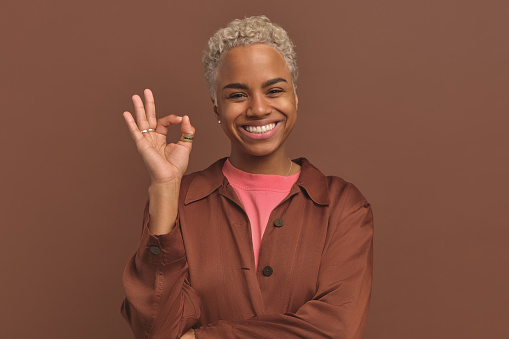 Young smiling African American woman with short blond hair makes OK gesture demonstrating answering question how are you doing or sending signal to start planned actions stands in brown studio