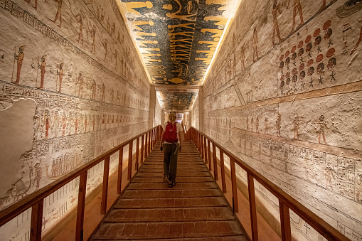 Valley of the Kings, Luxor, Egypt - July 26, 2022: Young woman walking in the tomb of Ramses V and Ramses VI also known as KV9. Tomb KV9 was originally constructed by Pharaoh Ramesses V. He was interred here, but his uncle, Ramesses VI, later reused the tomb as his own.

The tomb has some of the most diverse decoration in the Valley of the Kings. Its layout consists of a long corridor, divided by pilasters into several sections, leading to a pillared hall, from which a second long corridor descends to the burial chamber.
