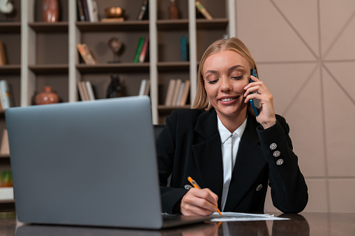 Smiling businesswoman take note and talk on the phone, laptop and document on desk. Office room with shelf and decoration on background. Concept of communication
