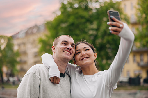 Beautiful young woman talking selfie with boyfriend outside in city. Happy young couple making a selfie with mobile phone outdoors.