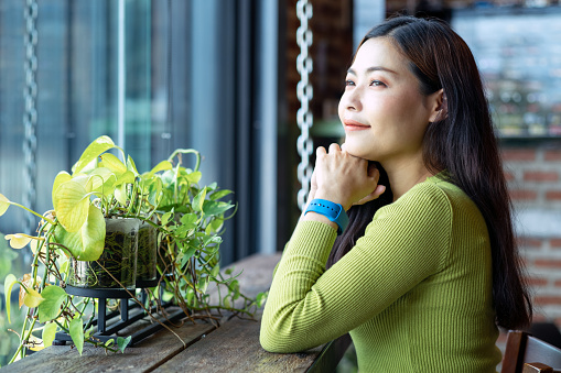 Asian beautiful young woman in green sweater sitting near cafe window and looking outside with copy space
