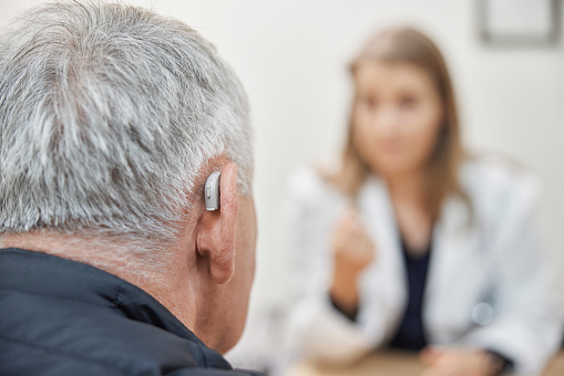 Close-up of a senior man with hearing aid on his ear visit his doctor at hearing clinic. Over the shoulder view of a elderly man wearing hearing machine sitting at medical clinic and listening to the female doctor advice.