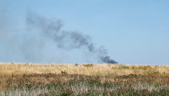 Black smoke rising into the sky above a parched grassland area as a wildfire burns in the hot summer climate.