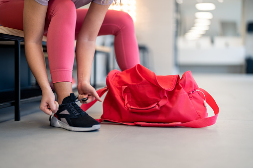 Cropped shot of fit sports woman in sportswear with gym bag wearing pink yoga pants and sneakers sitting on bench, getting ready for exercise session, tying her shoelaces in locker room at gym fitness studio.