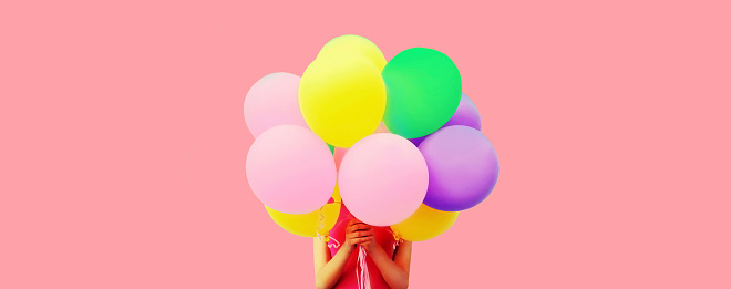Festive image of unknown woman covering her head and holding in hands bunch of colorful balloons on pink background