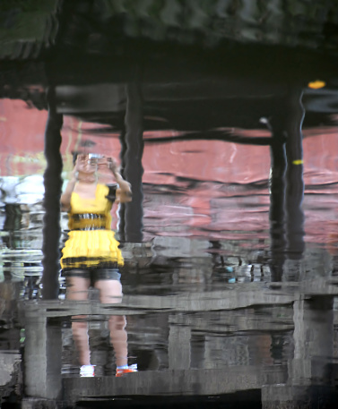 Water reflection of a woman in a yellow dress standing in a pavilion and taking a photo. Taken in the Yu Yuan Gardens in Shanghai, China.
