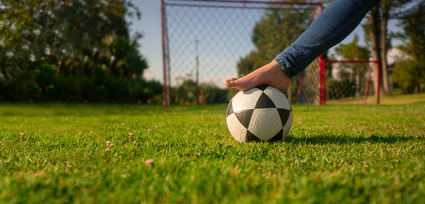 Close up on the leg of a woman in blue pants, barefoot in the middle of the park playing with a soccer ball with a soccer goal with nets and trees in the background during the morning of a sunny day